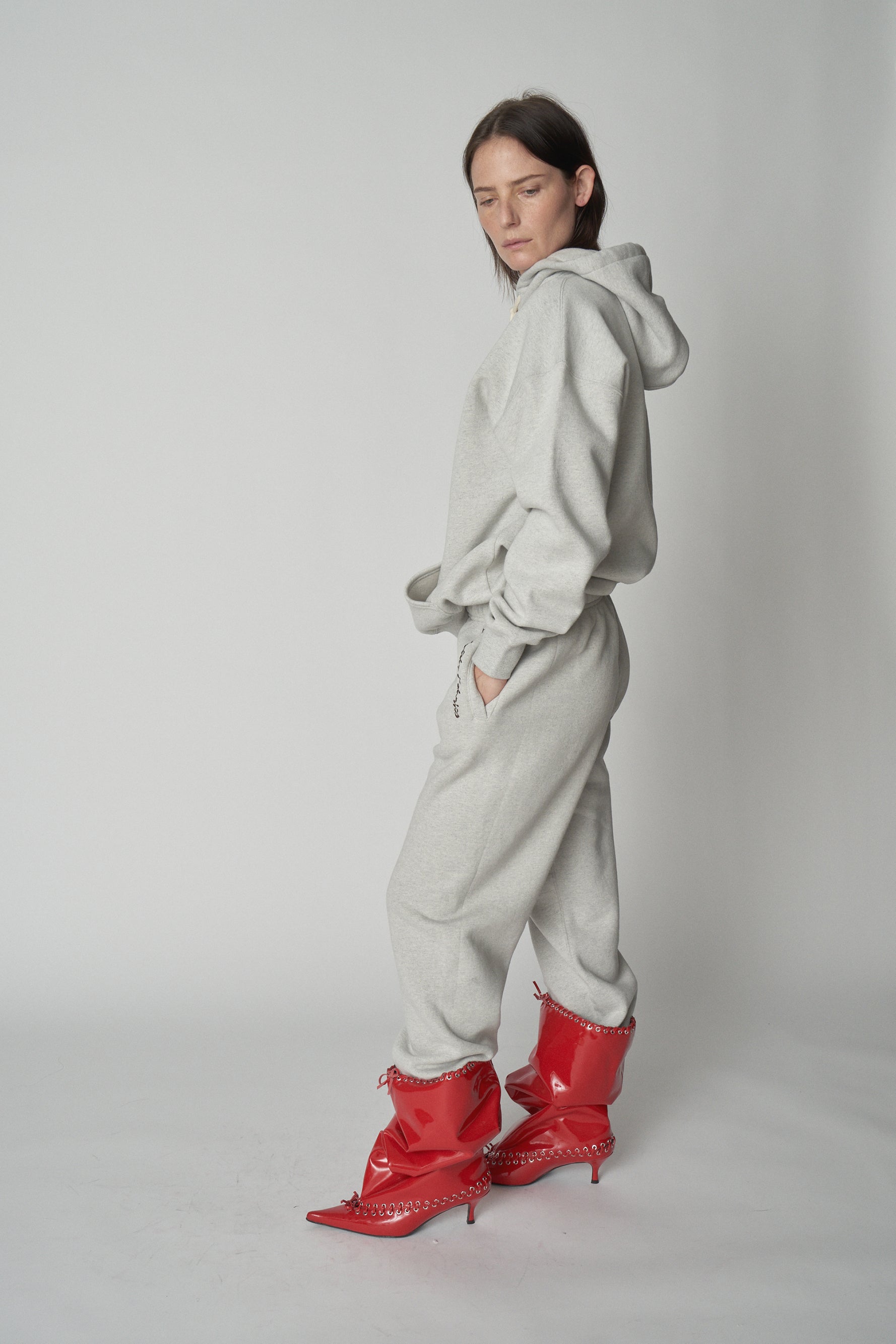 Full-length side view of model wearing grey Absolutely Fabrics hoodie and sweatpants with red heeled boots.