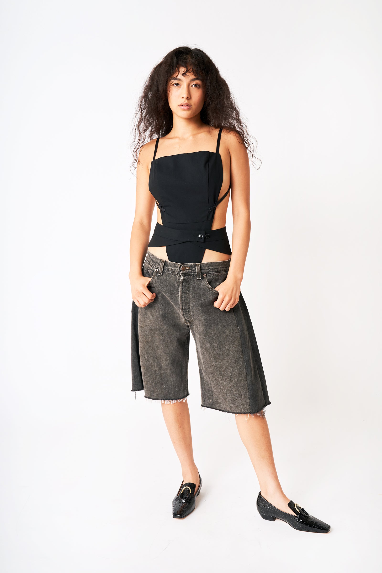 Full front view of model wearing black tank bodysuit with oversized black denim shorts and black loafers. 