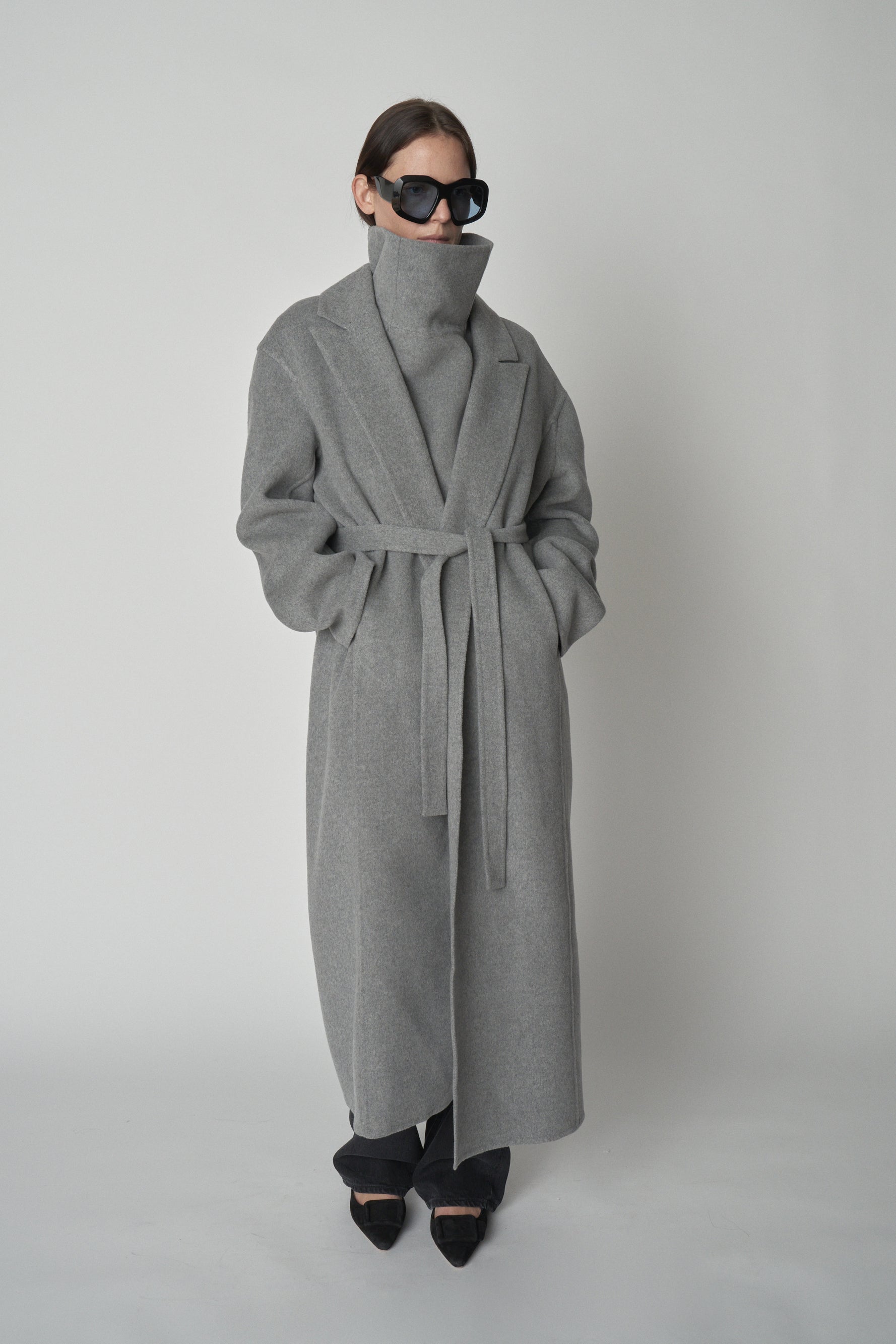 Front view of model wearing grey wool gillet and matching trench coat with large black sunglasses.